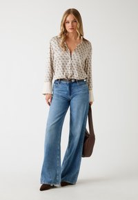 Blouse beige à motifs avec des manches longues, assortie à un jean en denim à jambes larges et des chaussures à talons marron. Tenant un sac à main marron foncé.