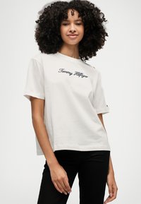 Jasno-beżowa bawełniana koszulka z krótkimi rękawami, z haftowanym granatowym logo "Tommy Hilfiger" i dekoracyjnymi guzikami na ramieniu.