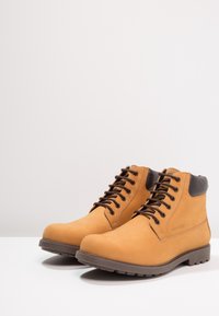 Bottes à la cheville en cuir tanné avec accents marron, bout rond, et fermeture à lacets noirs. Dotées d'une tige texturée et d'une semelle en caoutchouc durable.