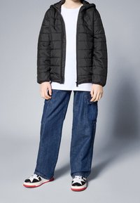 Chaqueta acolchada negra con cierre frontal y capucha, llevada sobre una camiseta blanca, combinada con unos pantalones cargo azules holgados y zapatillas negras con detalles rojos.