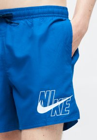Μπλε σορτς Nike με λευκό λογότυπο στο αριστερό πόδι, φορεμένα από άτομο με το χέρι στη δεξιά τσέπη, μπροστά σε απλό φωτεινό φόντο.