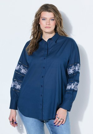 Mujer con cabello suelto y ondulado lleva una camisa azul marino con botones y bordado floral blanco en las mangas y vaqueros azul claro sobre un fondo sencillo.