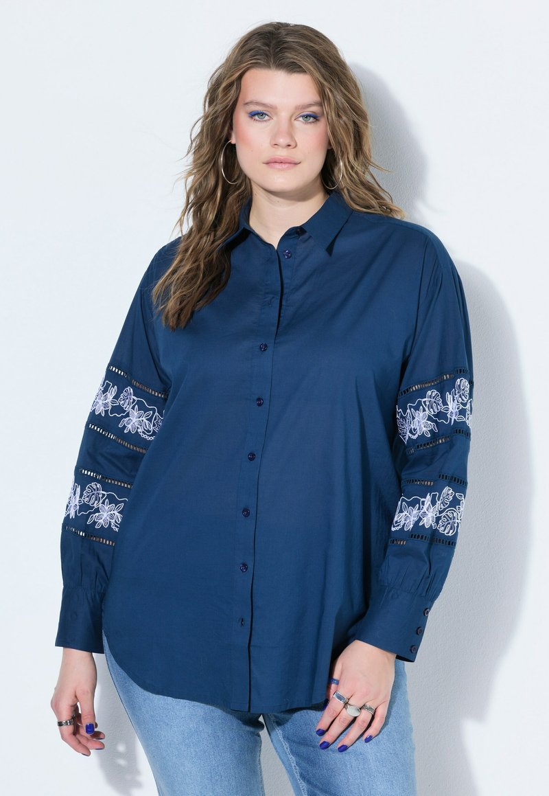 Femme aux cheveux lâchés et ondulés porte une chemise boutonnée bleu marine avec une broderie florale blanche sur les manches et un jean bleu clair sur un fond uni.