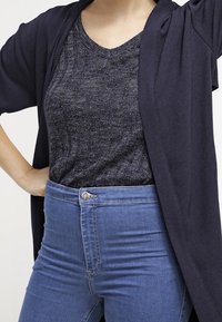 Marine open cardigan over een donkere, gestructureerde top, gecombineerd met high-waisted blauwe jeans. Cardigan heeft brede mouwen en een losse pasvorm.
