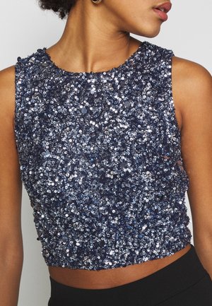 Femme portant un crop top bleu marine sans manches, densément couvert de sequins brillants, associé à un bas noir.