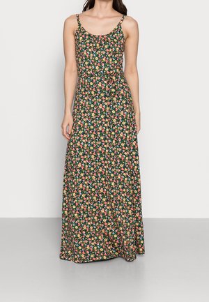 Femme portant une robe longue fleurie sans manches avec de fines bretelles et une petite ceinture nouée à la taille, debout devant un fond uni.