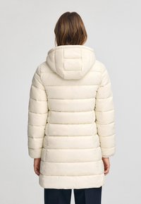 Puffer jakke i lys creme med kviltet design og fastgjort hætte. Har en mellemlang snit og lange ærmer. Glat, vandafvisende tekstur.