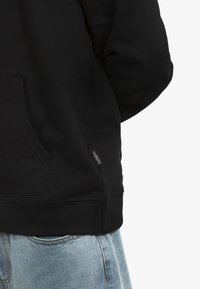 Felpa nera con tasca a canguro e orlo a costine. Presenta un piccolo etichetta tessuta sul lato inferiore. Abbinata a jeans in denim azzurro chiaro.