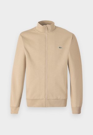 Šviesiai rudos spalvos užtraukiamas džemperis su stojančiu apykakle, minkšta medžiaga, raštuotos rankogaliai ir mažas žalias Lacoste logotipas ant krūtinės. Paprastas ir minimalistinis dizainas.