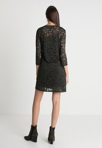 Svart spetsklänning med trekvartsärmar, blommönster, figursydd silhuett och vågig nederkant, tillsammans med mörka klackiga ankelskor.