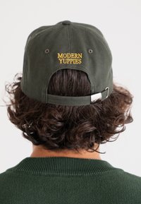 Dark green baseball cap with embroidered yellow text "MODERN YUPPIES" on the back. Adjustable strap with metal clasp visible.