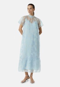 Jasnoniebieska sukienka w kwiatowy wzór z przezroczystą warstwą, krótkimi rękawami i warstwowym dołem. Połączona z srebrnymi sandałkami na obcasie.