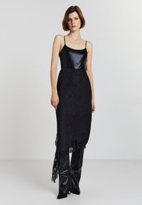 Top di raso nero con spalline sottili abbinato a una gonna nera con dettagli in pizzo. Presenta un lungo orlo asimmetrico e pantaloni lucidi simili a pelle.