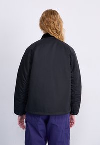 Schwarze isolierte Jacke mit glatter Oberfläche, Raglanärmeln und einem kontrastierenden Kragen. Sichtbare Nähte entlang der Seiten und Ärmel.