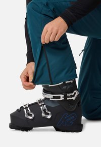 Persona inginocchiata, mentre slaccia i pantaloni da sci blu sopra uno scarpone da sci K2 nero con fibbie argento su sfondo bianco.