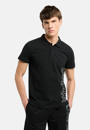 Jonge man draagt een zwart poloshirt met tekst aan de zijkant en zwarte broek, staat met handen in de zakken tegen een effen achtergrond.