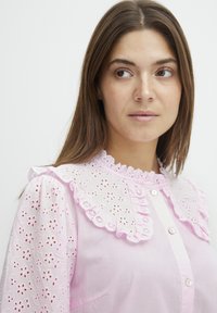 Svetloružová blúzka s vyšívaným bielym golierom, balónovými rukávmi a predným zapínaním na gombíky. Textúrovaná látka má vzory s vyrezávanými kvetmi.