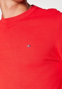 T-shirt rouge à col rond en coton. Présente un petit logo bleu et blanc sur la poitrine. Manches courtes et coupe ajustée.