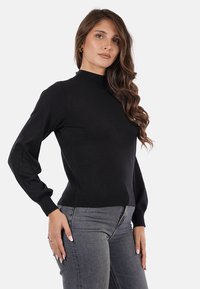 Maglione nero a maglia con colletto alto e maniche lunghe, caratterizzato da una silhouette aderente e polsini a coste, abbinato a jeans grigio scuro.