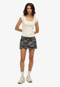 Roomkleurige geribbelde top met gegolfde randen, gecombineerd met een grijze camouflagerok met cargozakken. Casual sneakers en witte sokken maken de look af.