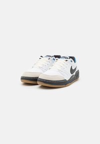 Zapatillas Nike blancas con una puntera texturizada en gris, detalles negros en los lados y una suela de goma color caramelo. Presenta detalles azules en la lengüeta.