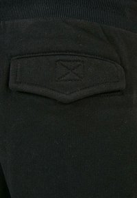 Fekete szövet zseb, varrott fedéllel, amelyen egy X-alakú varrás található egy sötét ruházati terméken.
