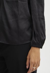Schwarze, leichte Bluse mit glatter Textur, gerafften Ärmeln und lockerer Passform; getragen mit schwarzen Hosen und einem schlichten silbernen Ring.