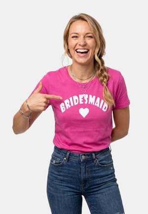 Mujer sonriente con cabello rubio trenzado lleva una camiseta rosa que dice "BRIDESMAID" con un corazón, señalando el texto.