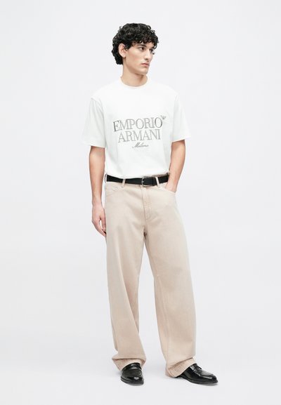 Biele bavlnené tričko so sivým logom "Emporio Armani Milano", kombinované s béžovými širokými nohavicami a čiernymi koženými topánkami.