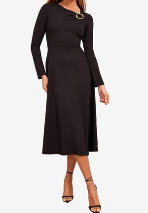 Czarna sukienka z długim rękawem, z dopasowaną górą i rozkloszowanym dołem, ozdobiona charakterystyczną okrągłą broszką przy dekolcie. Gładka tekstura.