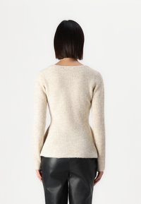 Un pull tricoté beige clair avec des manches longues, texture côtelée et taille évasée, porté sur un pantalon en cuir noir, vu de dos.