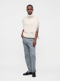 Femme portant un pull sans manches à col roulé blanc avec des franges et un jean bleu clair, posant avec une main dans la poche et des chaussures noires.
