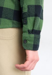 Main reposant sur le côté, portant une chemise à carreaux verts et noirs à manches longues avec des boutons au poignet et un pantalon beige avec une poche visible.