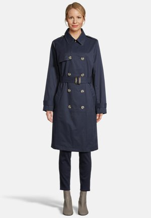 Vrouw draagt een marineblauwe dubbelgeknoopte trenchcoat met riem, donkere broek en grijze enkellaarzen, staand tegen een effen witte achtergrond.