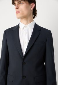 Tumes sinine bleiser kahe nööpiga, noaga krae ja rinna taskuga, koos valge kraejoonega särgiga. Sujuv tekstuur, kohandatud lõige.