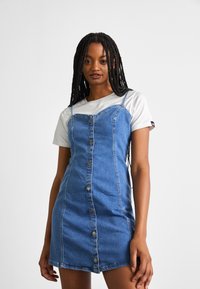 Denim overall dress in medium blue with a fitted shape, button-down front, and thin shoulder straps. Paired with a white short-sleeve shirt.
