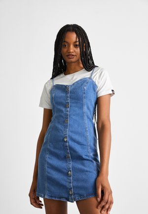 Denim overall dress in medium blue with a fitted shape, button-down front, and thin shoulder straps. Paired with a white short-sleeve shirt.