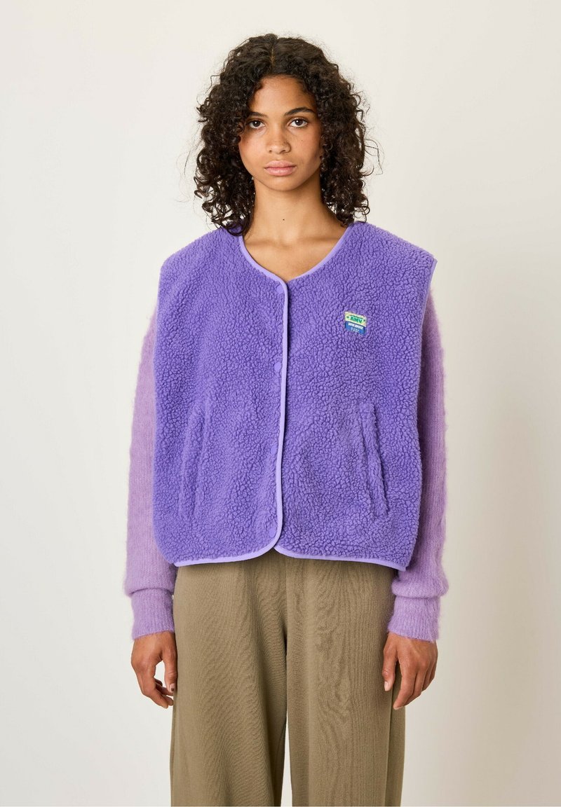 Paarse fleece vest met ronde halslijn, voorzakken en lavendelaccenten. Draagt boven een lichtpaars sweater met textuur in de mouwen.