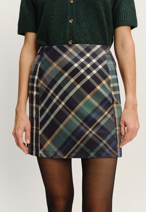 Kobieta w zielonym swetrze na krótki rękaw z guzikami, w kratkowanej mini spódnicy w odcieniach granatu, zieleni i beżu oraz czarnych przezroczystych rajstopach.