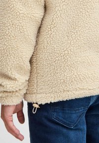 Beige fleece jacket with a textured, fluffy surface. Features a cinch drawstring at the hem, paired with dark blue denim jeans.