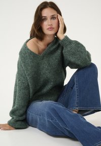 Pull vert en maille tricotée avec un large col en V et une coupe surdimensionnée, associé à un jean bleu évasé. Tissu texturé, silhouette décontractée.