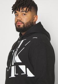 Black hoodie featuring large white graphic prints, drawstring hood, and soft fabric. Design includes distinct shapes and bold lettering.
