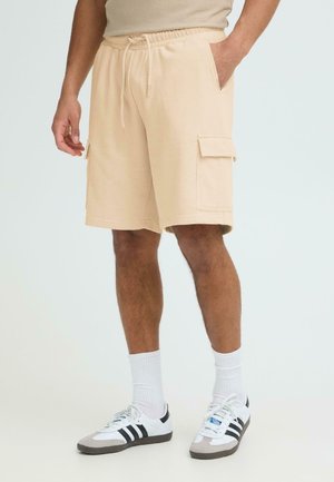 Moški v bež cargo kratkih hlačah z žepi, belih nogavicah s kratkim robom in belih supergah s črnimi črtami, stoji na svetlem ozadju.