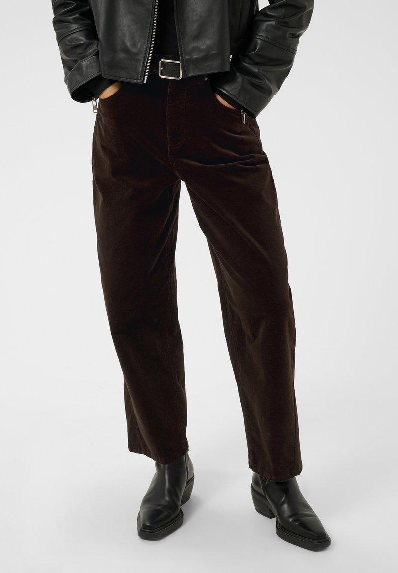 Personne portant un pantalon en velours côtelé brun foncé, une veste en cuir noire, une ceinture noire et des bottines en cuir noir aux chevilles, les mains dans les poches.