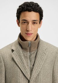 Brown houndstooth blazer with a high-knit collar, featuring a zipped inner layer. The material appears textured, combining comfort and structure.