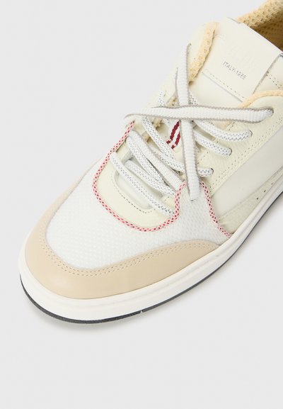 Sneaker bianca e beige realizzata con materiali in rete e pelle. Presenta accenti grigi e rossi, lacci rotondi e una suola in gomma testurizzata.