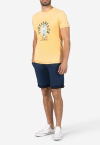 Yellow short-sleeve t-shirt with graphic print, paired with navy shorts. White sneakers complete the outfit, featuring rolled hems.