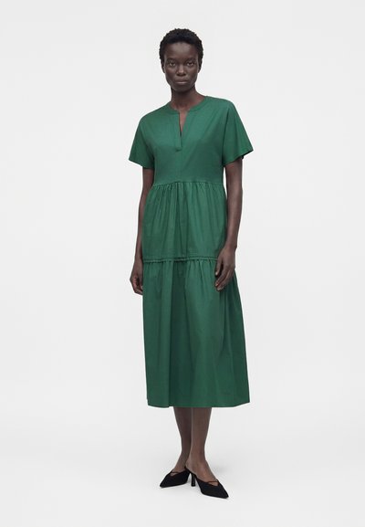 Γυναίκα όρθια, φορόντας πράσινο μίντι φόρεμα με κοντά μανίκια και πτυχώσεις, μαζί με μαύρα παπούτσια με χαμηλό τακούνι, μπροστά σε λευκό φόντο.