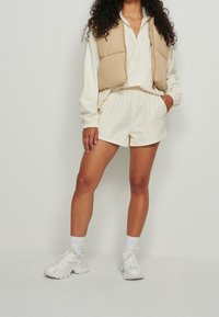 Beżowa pikowana kamizelka na białej koszuli z długim rękawem, w połączeniu z białymi szortami i białymi tenisówkami. Charakteryzuje się luźnym krojem i elastycznymi mankietami.