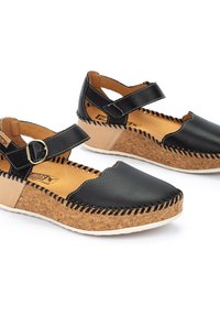 Sandalias de cuero negro con suelas de corcho en forma de cuña, que presentan una correa con hebilla lateral y costura decorativa a lo largo de los bordes. Textura de superficie suave.